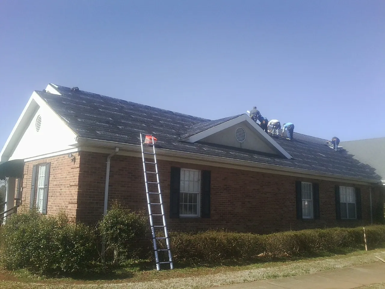 Crew tearing shingles off a brick ranch roof with a ladder set — Emergency Roof Repair work by our crew in East Islip