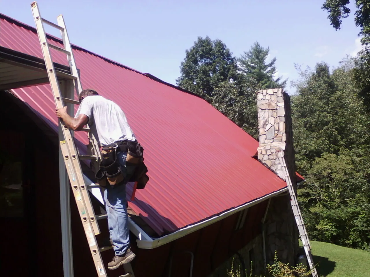 Roofer on a ladder beside a red metal roof and stone chimney — Soffit & Fascia Repair by Roofing Brothers in East Islip